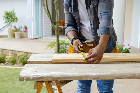 African American male measuring plank using tape marking pencil on patio wearing tool belt