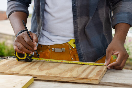 African American man measuring plywood with yellow tape measure on bench, wearing tool belt