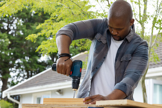 African male using cordless drill on wooden plank on sawhorse in house backyard, wearing gray shirt