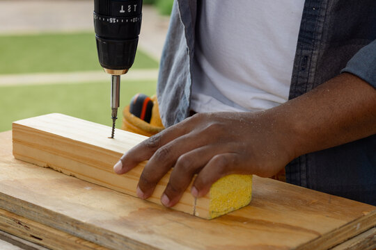 African American man holding pine board as driving screw with drill on patio in denim shirt