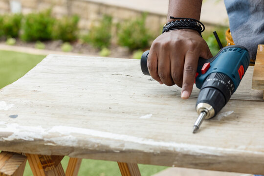African American man using drill drilling plank on sawhorses in yard with bracelets, copy space