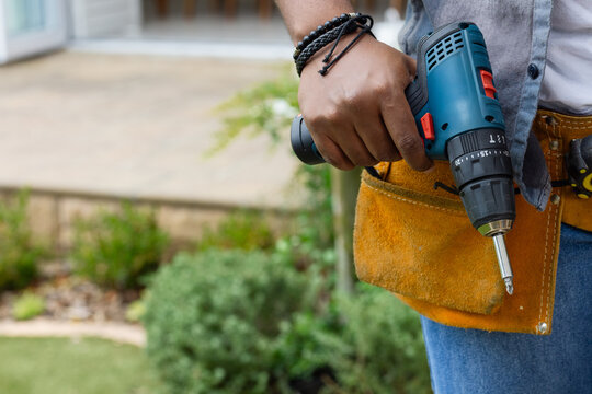 African man holding blue cordless drill, wearing grey shirt and tool belt at patio, copy space