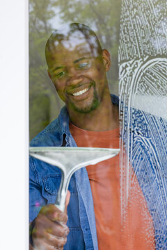 African man cleaning large glass window at home with hand squeegee, smiling in denim jacket