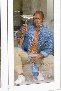 African man kneeling, cleaning glass door at home in denim jacket with squeegee and blue bottle