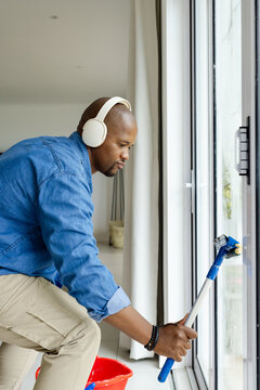 Man kneeling and cleaning sliding glass door at home with squeegee, bucket, denim shirt, headset