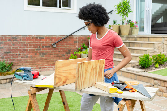 African woman DIYer working on plywood at house patio sawhorse wearing red shirt, goggles, sander