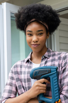 Woman in 20s standing in doorway holding blue cordless power nailer, wearing checkered shirt
