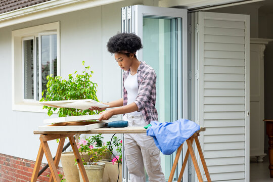 Inspecting wooden boards in plaid shirt and cargo pants at house threshold, with drill and plants