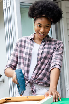 African American woman holding blue staple gun steadying wooden frame at porch wearing plaid shirt