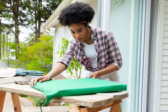 Stretching woman in pink plaid smoothing green fabric over foam pad on porch workbench, blue tool