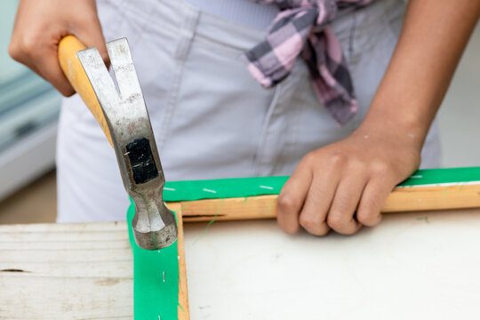 Woman wearing plaid shirt securing green fabric to wooden frame with yellow claw hammer at bench
