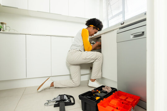 African woman kneeling on kitchen floor, working under sink with yellow shirt and toolbox