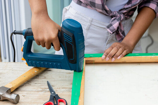 African American woman wearing pink plaid shirt using blue staple gun steadying frame on workbench