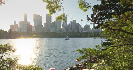 Obraz premium Australia, Sydney city: warm sunset over Sydney Harbour framed by lush green trees, light reflecting on calm water, with boat sailing cityscape skyline. Drone flight in forest park window