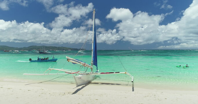 Turquoise tropical sea washing White Beach on Boracay Island, Philippines, with a paraw sailboat resting on the shore and a motorboat carrying tourists in the background