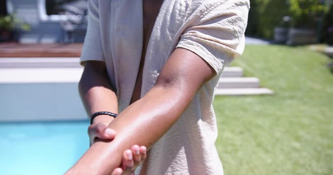 Vertical video: Rubbing man smoothing right forearm at backyard pool, wearing beige shirt wristband