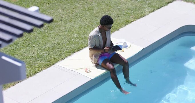 African American man sitting on pool edge, reading tablet and dipping feet to cool
