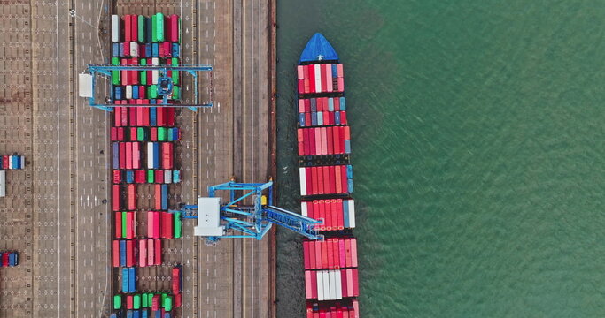 Cargo ship docks at a busy industrial port, having colorful shipping containers loaded and unloaded by cranes, representing global logistics and transportation. Top down aerial drone flight