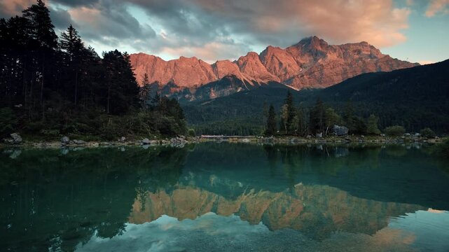 Mountains lit with warm sunset light over a scenic Lake. Panning shot of a painterly scenery in Bavaria, Germany, with teal calm water reflecting the landscape.