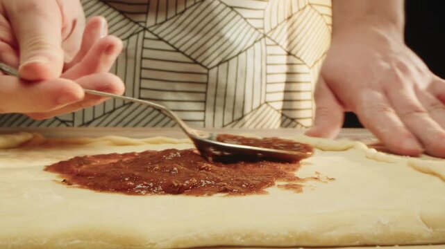 Close up of spoon spreading rich tomato sauce over raw dough. Pizzeria chef in patterned apron executing fast smooth motion applying sauce to pizza dough on paper in kitchen. Cooking an Italian dish