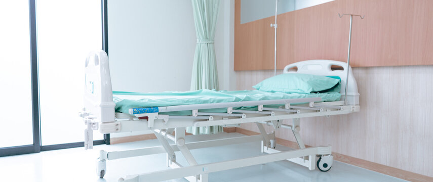A hospital room shows an empty bed ready for a patient. The bed has light blue sheets and there are some medical devices on the wall. Bright light comes through the window.