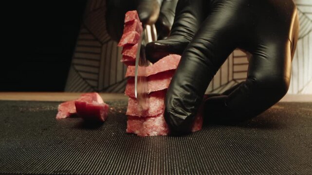 Close up of male gloved hands slicing smoked salami into cubes for gourmet prep. Souschef in apron and prep cook techniques with sharp knife on cutting board. Unhealthy diets and trans fats