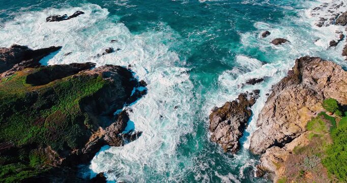 Top view of rocky inlet with crashing waves. Aerial shot of an ocean inlet with green vegetation on rocky cliffs and white surf in Big Sur, California.