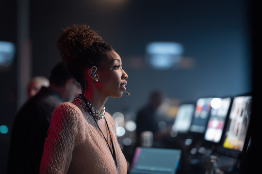 Medium view of a Black woman TV presenter or producer wearing headset with mic, standing backstage. Out-of-focus screens and active crew in background. Broadcasting environment. Media expert.