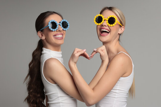 Studio shot of two young women best friends wearing funny party glasses laughing and making heart shape gesture on gray background