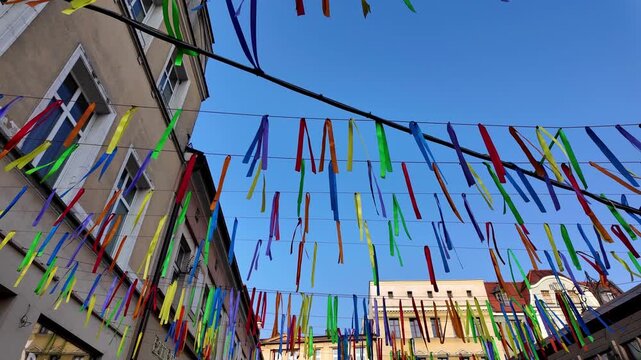 Colorful ribbons hanging over a city street between old buildings. Joyful festival decoration against a clear blue sky, creating a festive atmosphere during a local cultural event or parade.
