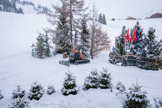 A red and gray snow groomer sits by fir and larch trees, fresh tracks mark a ski slope, flagpoles with national flags stand near chalets in Oberlech, Austria, snowfall
