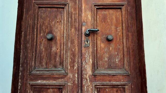 Vintage brown wooden door with peeling paint, brass handle and ornate metal fittings. Close-up detail of an old entrance showing wood texture, history, and architectural patina of time.