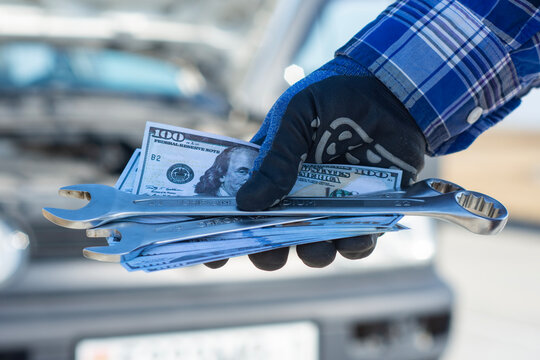Close up of mechanic hand holding wrenches and dollar cash near car, representing vehicle repair cost and service pricing. Automotive maintenance, financial expense and auto service business concept.