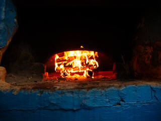 Bright orange flames consume firewood logs inside dark arched chamber of rustic blue painted stone oven during traditional baking process. © Dmitry