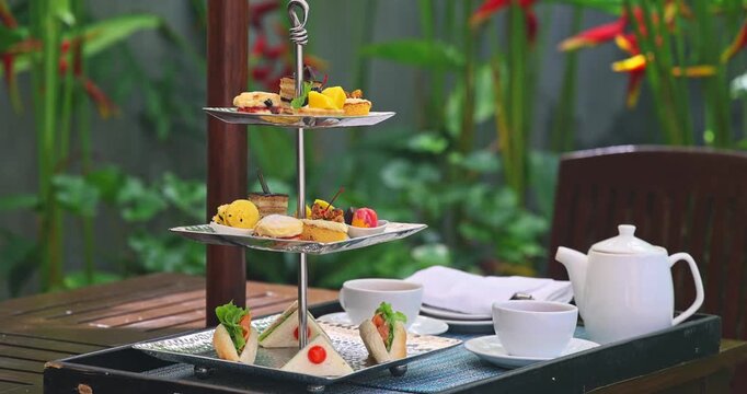 Three-tier afternoon tea stand featuring sandwiches and pastries sitting on wooden table in tropical garden, bright natural light, luxury hospitality and leisure lifestyle.