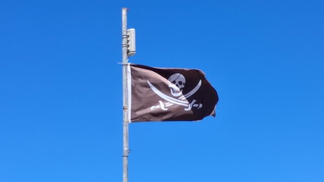 Close-up on a pirate flag with skull and crossed swords fluttering in wind against clear blue sky.