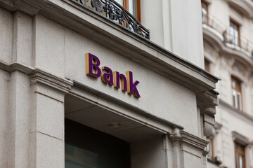 Obraz premium Urban bank entrance with purple sign on grey stone facade and decorative balcony detail in a european city street showing commercial building frontage