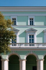 Naklejka premium Pale mint neoclassical facade with arched ground floor colonnade, ornamental pediments over windows, balustrade balcony and leafy tree framing the elegant exterior