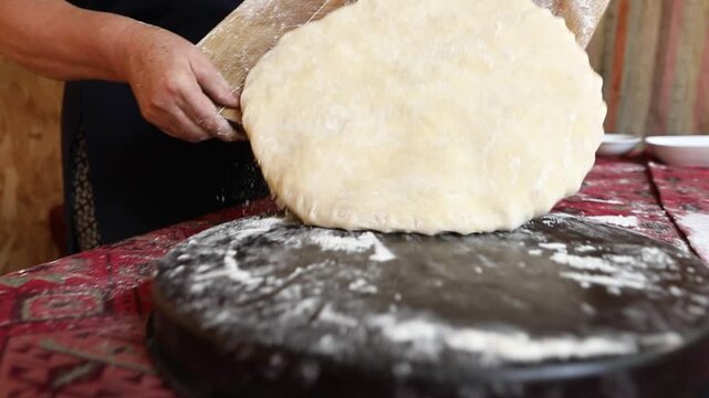 A woman in a traditional apron carefully slides a thick, round piece of raw dough from a floured wooden board into a dark baking pan on a table.

