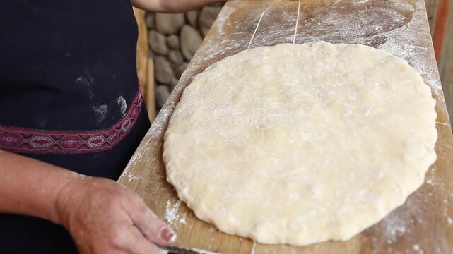 A woman in a traditional apron carefully slides a thick, round piece of raw dough from a floured wooden board into a dark baking pan on a table.

