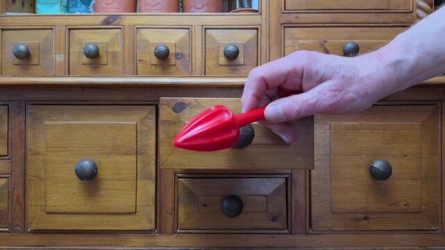 A senior man&rsquo;s hand opening and closing a drawer in an old pine kitchen dresser, to remove a red plastic, manual juicer too, to remove juice from citrus fruits while trapping pips and pulp.