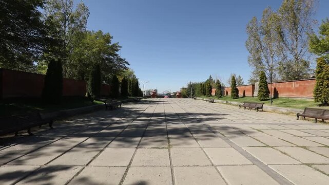 The camera moves slowly along a paved pathway lined with trees and benches, eventually passing through a large, open wrought-iron gate to reveal a bright, sunlit park entrance.