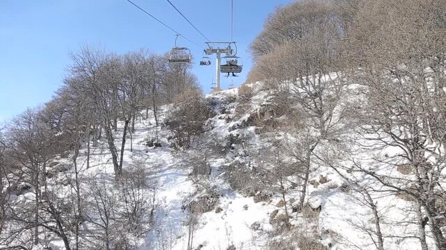 A scenic point of view shot from a ski chairlift descending a snow-covered mountain, passing bare trees, with majestic snow-capped peaks and a valley in the hazy distance.