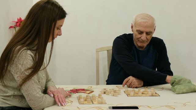 Granddaughter and a senior grandfather spending quality time together, playing an educational wooden board game, which helps develop logic, memory, and cognitive skills for the elderly at home