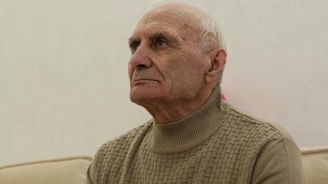 Pensive senior adult man with white hair sitting alone on a couch, looking away with a serious and worried expression, reflecting on his life memories and feeling loneliness