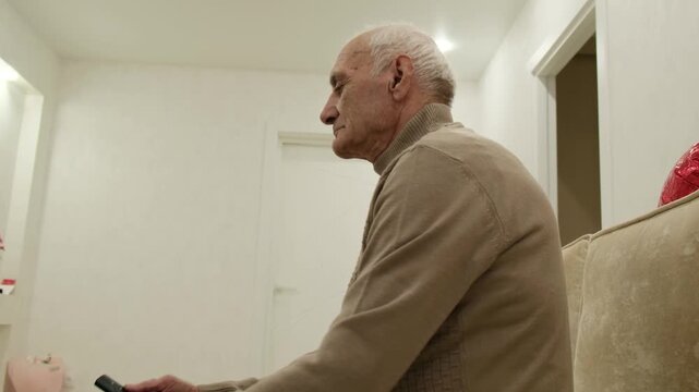 Elderly man resting on a comfortable sofa in his living room, attentively watching television and using a remote control for zapping through different channels for entertainment