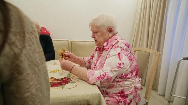 Group of senior friends enjoying a card game together while sitting around a table in a cozy room, focusing on a woman sorting her hand, highlighting social activity and leisure for elderly people
