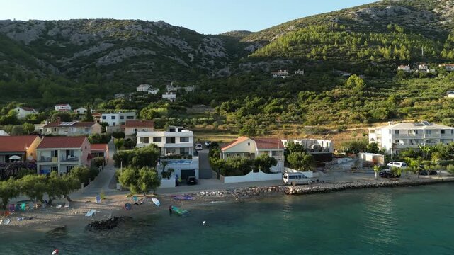Aerial drone view of a modern white Mediterranean villa with a private pool at golden hour, surrounded by a hillside coastal neighborhood with lush greenery and terracotta roofs.