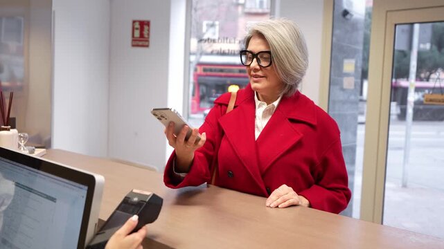 Stylish mature woman with grey hair in a red coat using her mobile phone for a contactless payment at a dental clinic reception desk