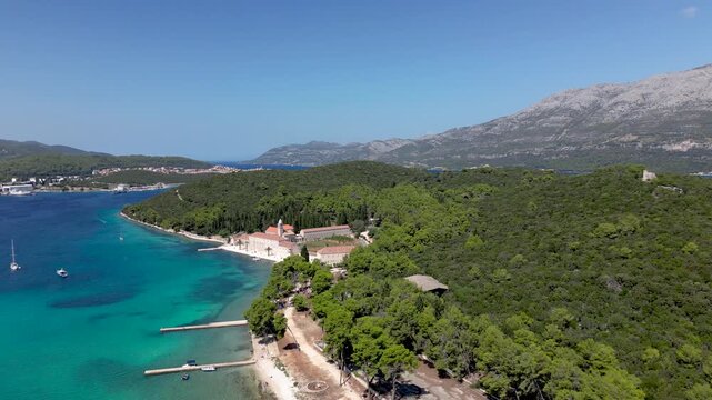 Cinematic drone aerial over Badija Island, Croatia, gliding along a pine forest shoreline and pebble beach with a coastal path and dramatic mountains in the background.